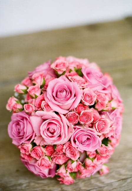 A round bouquet arrangement of fresh pink roses with tightly coiled petals, accompanied by smaller rosebuds and green foliage, sits on a rustic wooden surface. The roses vary in shade from soft pastel