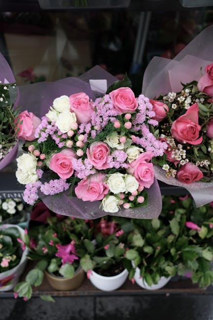 A bouquet of fresh pink roses and white carnations, accented with delicate light purple statice, arranged with lush green foliage. The flowers are expertly wrapped in pale purple tissue paper, creatin