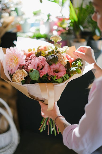 A person holding a floral bouquet arranged with pink peonies, white and purple blossoms, and greenery, wrapped in cream-colored paper with a beige ribbon. The bouquet is being prepared for flower deli