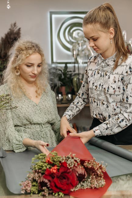 Two women are engaged in preparing a floral gift, with one woman, dressed casually in a patterned blouse, holding a bouquet of fresh red roses, pink carnations, and other small flowers wrapped in red 
