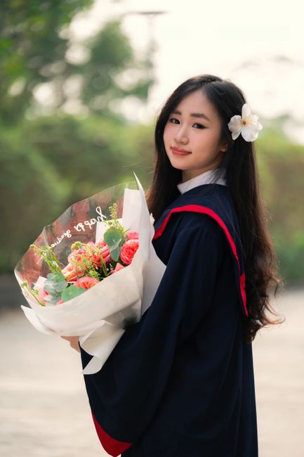 A young woman with long dark hair, wearing a navy blue graduation gown with a red trim and a white shirt underneath, is holding a large bouquet of fresh flowers. The bouquet is wrapped in white and tr