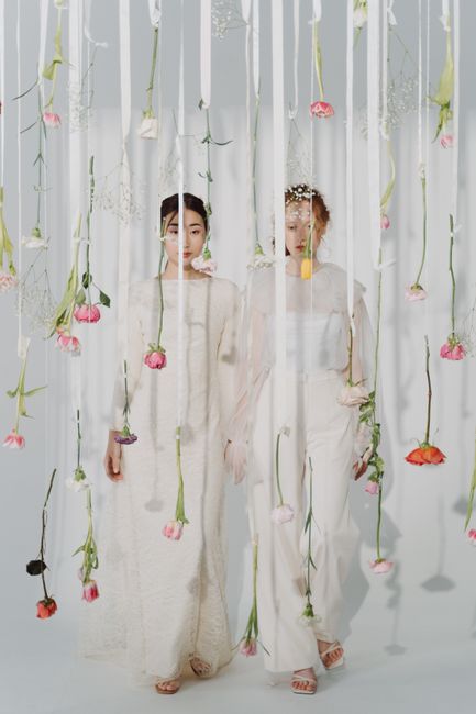 Two women dressed in elegant white gowns stand amidst hanging floral arrangements of pink and peach roses with green stems, suspended from the ceiling by thin, nearly invisible strings. The women, wit