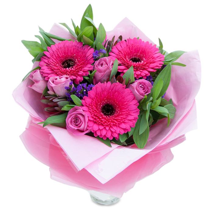 I picture this bouquet as a short, fragrant ode to spring: three fuchsia Gerbera daisies open wide, their radiant petals forming a bright, graphic focal point around dark centres that give the composition contrast and structure. Gentle, dusty-rose and lavender-pink roses nestle in between, their petals still forming tight, graceful spirals that suggest tenderness and newness. Sprays of tiny purple statice add a whisper of wildflower charm, their delicate clusters balancing the larger blooms and lending textural interest. A generous sweep of verdant foliage-broad, glossy leaves like salal and slender eucalyptus - creates a garden-like backdrop that enhances the saturation of the pinks and anchors the bouquet with cool green. The flowers are wrapped with an inner layer of pale pink matte tissue, soft to the touch, and an outer sheet of translucent, slightly crinkled fuchsia-pink cellophane that shimmers faintly and unifies the colour story. You can glimpse the top of a clear glass vase beneath the wrapping, a quiet cue that this piece is meant to be displayed immediately, whether as a bedside surprise or a centrepiece at a small gathering. Under soft daylight the textures read clearly: velvety rose petals, crisp gerbera rays, fragile statice stems. The scent is subtle - rose perfume brightened by a clean green note - and the overall mood is uplifting and intimate. It's the kind of bouquet a neighbour might send across Egham or to a student at Royal Holloway to mark a birthday, a thoughtful thank-you, or a gentle celebration.